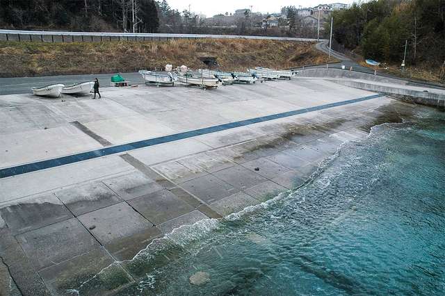 Рибний порт в районі Кусакідзава, де зупинили реалізацію плану будівництва хвилезахисних стіни вартістю 1,28 млрд ієн. На задньому плані видно насип колишньої залізничної лінії JR Кесеннума. Тепер це дорога для автобусного сполучення 